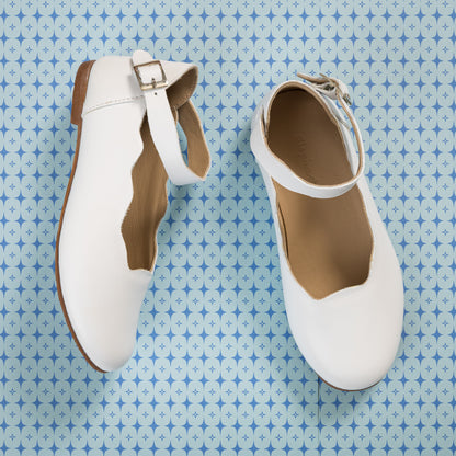 White children's ballerina flats with scalloped edges and ankle buckle strap on brown sole.