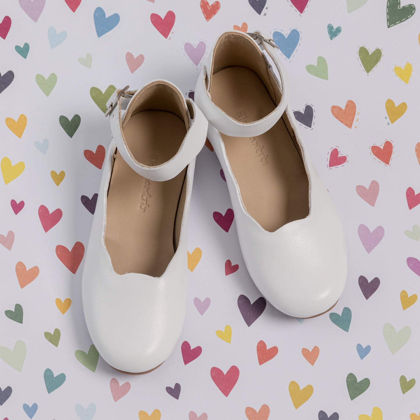 White toddler ballerina flats with scalloped edges and ankle straps on colorful heart pattern background.