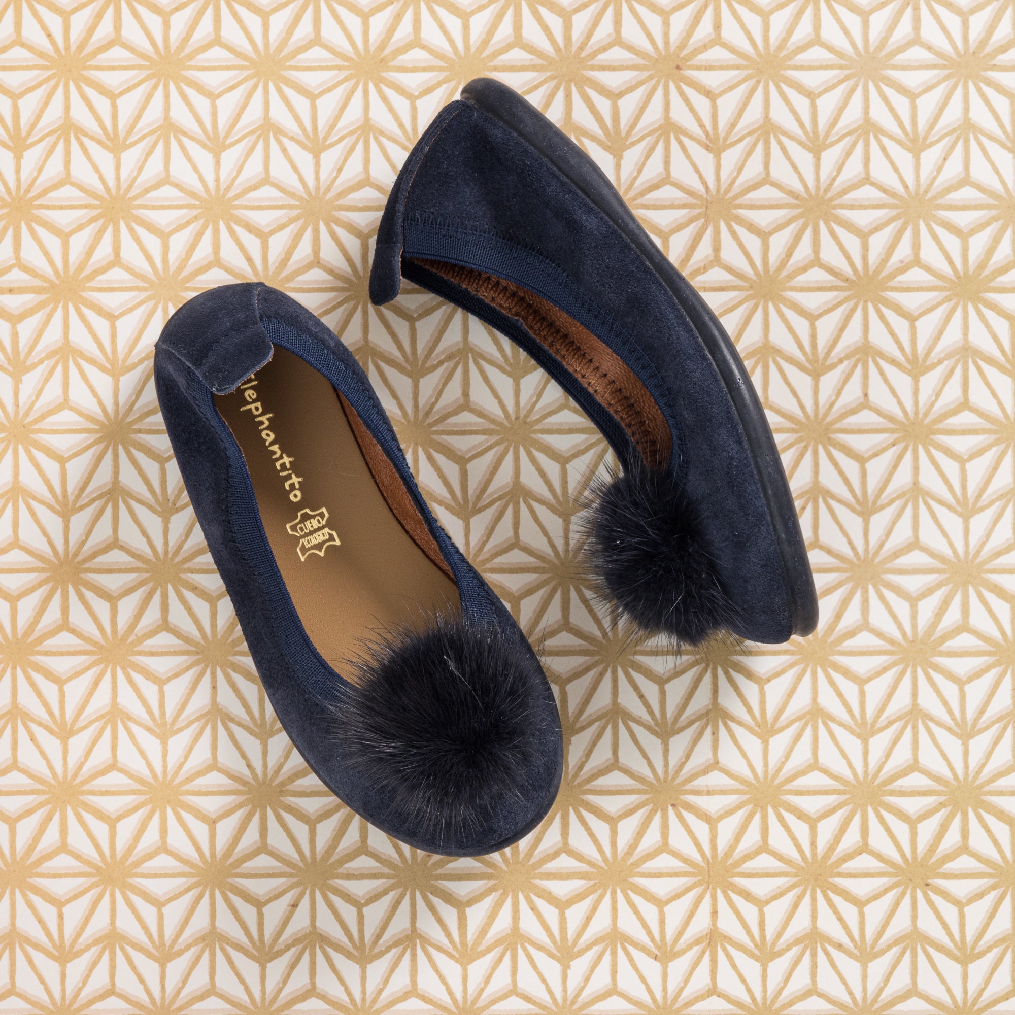 Blue suede ballerina flats with black pom poms on toes and brown insoles on geometric patterned background.