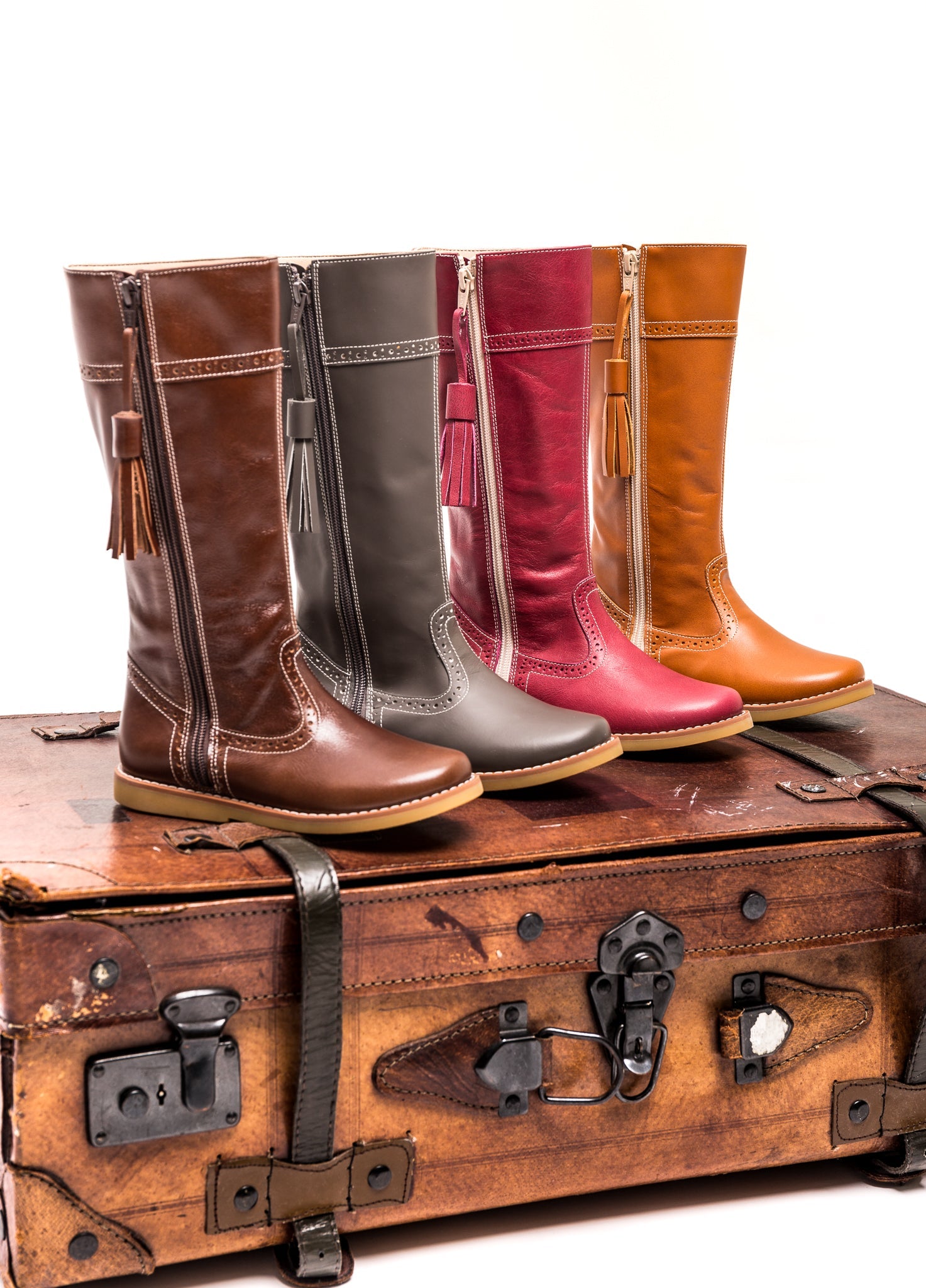 Brown children’s riding boots with tassel detail, rounded toe, and contrast stitching, arranged on vintage suitcase.