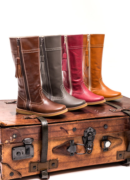 Brown children’s riding boots with tassel detail, rounded toe, and contrast stitching, arranged on vintage suitcase.