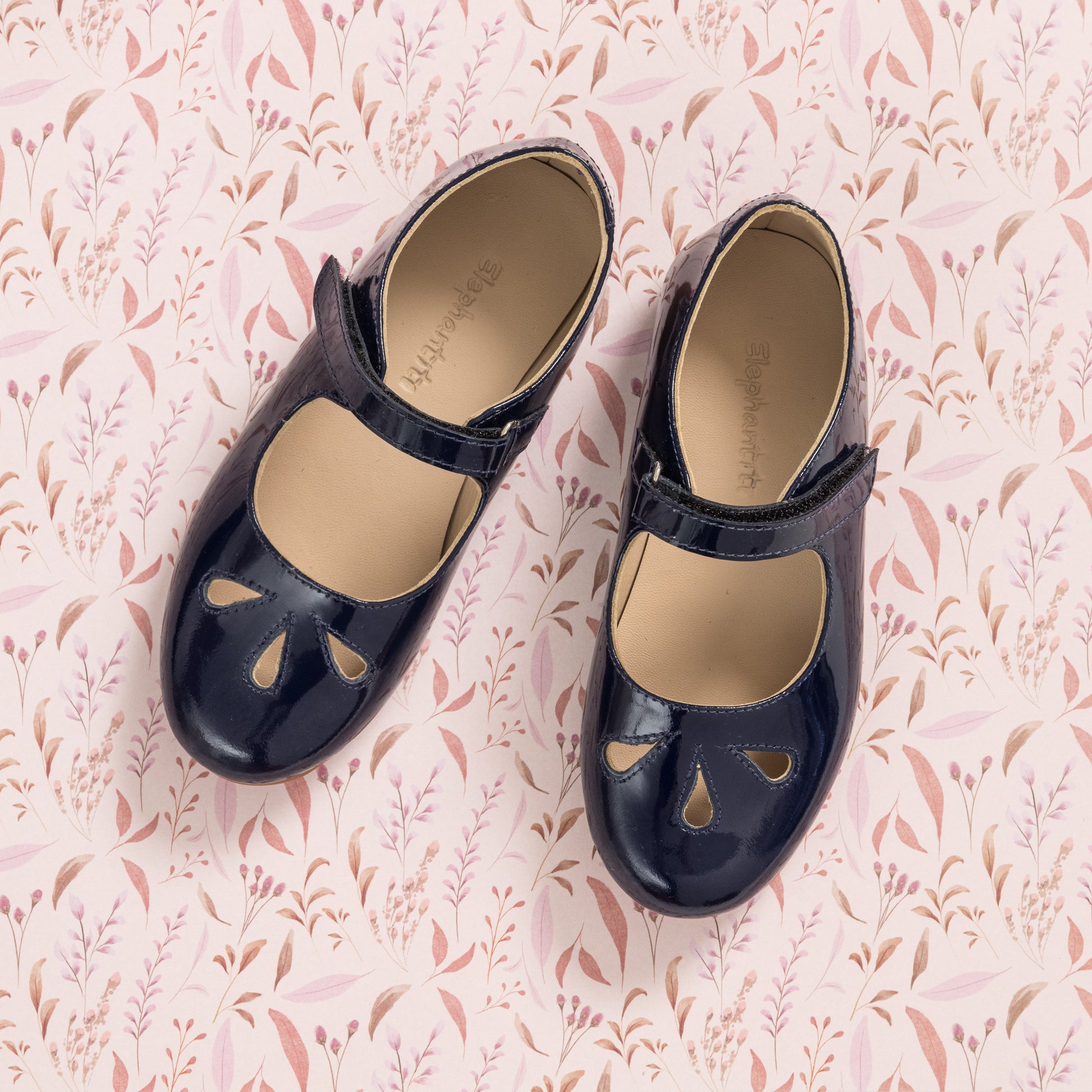 Navy blue patent leather girls Mary Jane shoes with teardrop cutouts and strap closure on pink floral background.