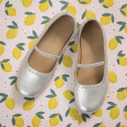 Silver metallic toddler ballerina flats with a thin strap on a lemon-patterned background.