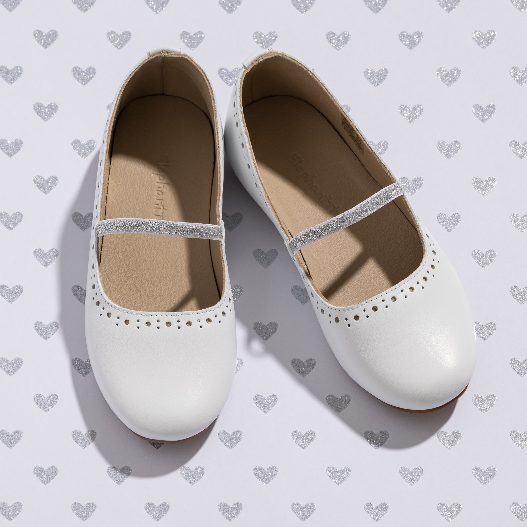 White toddler ballerina flats with perforated details and glittery strap on heart-patterned background.