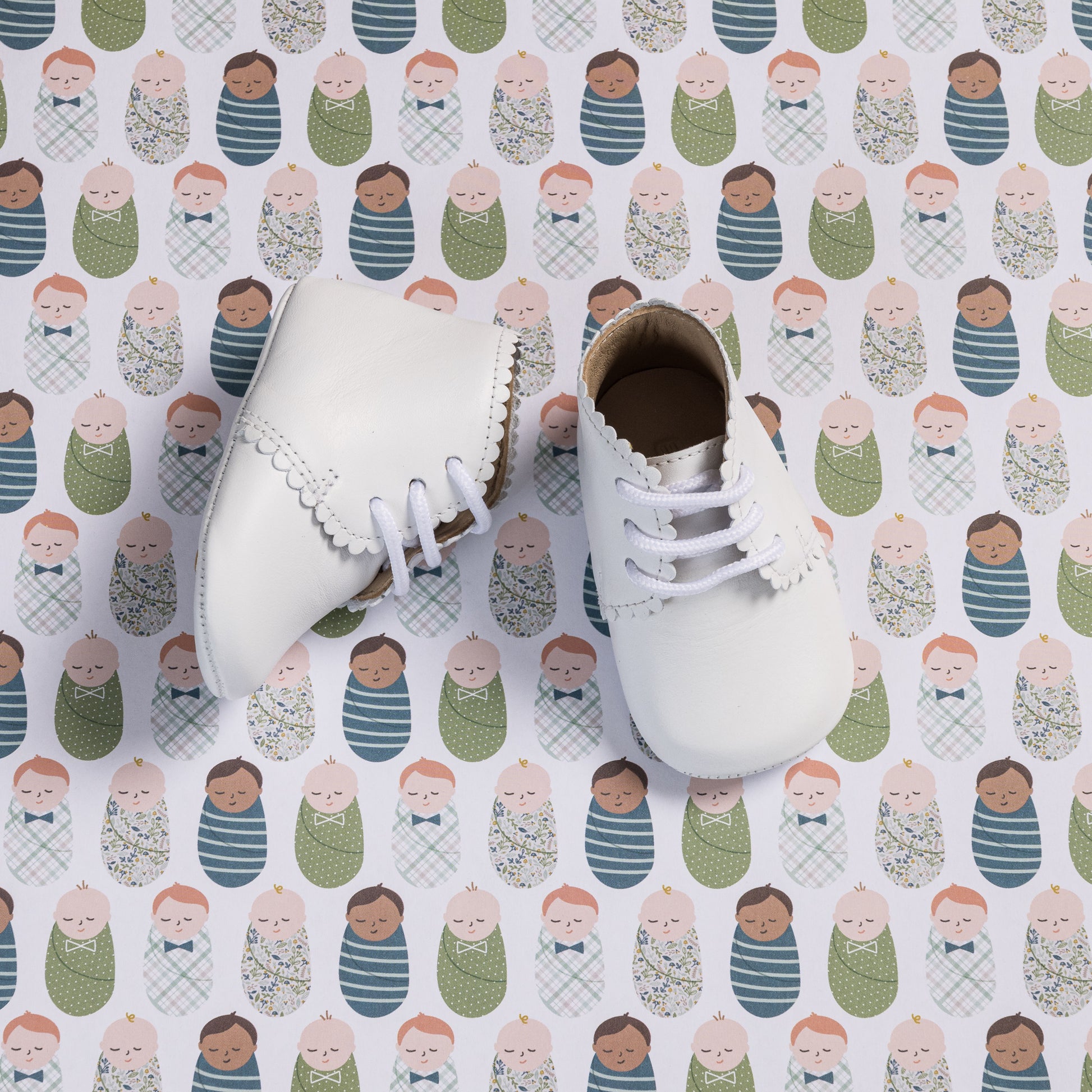 White leather baby booties with scalloped edges and white laces on patterned background.