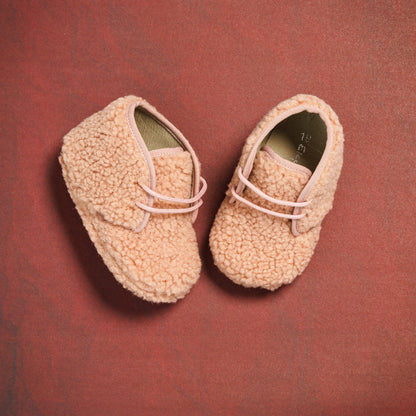 Pink shearling baby booties with soft textured exterior and elastic laces on red background.