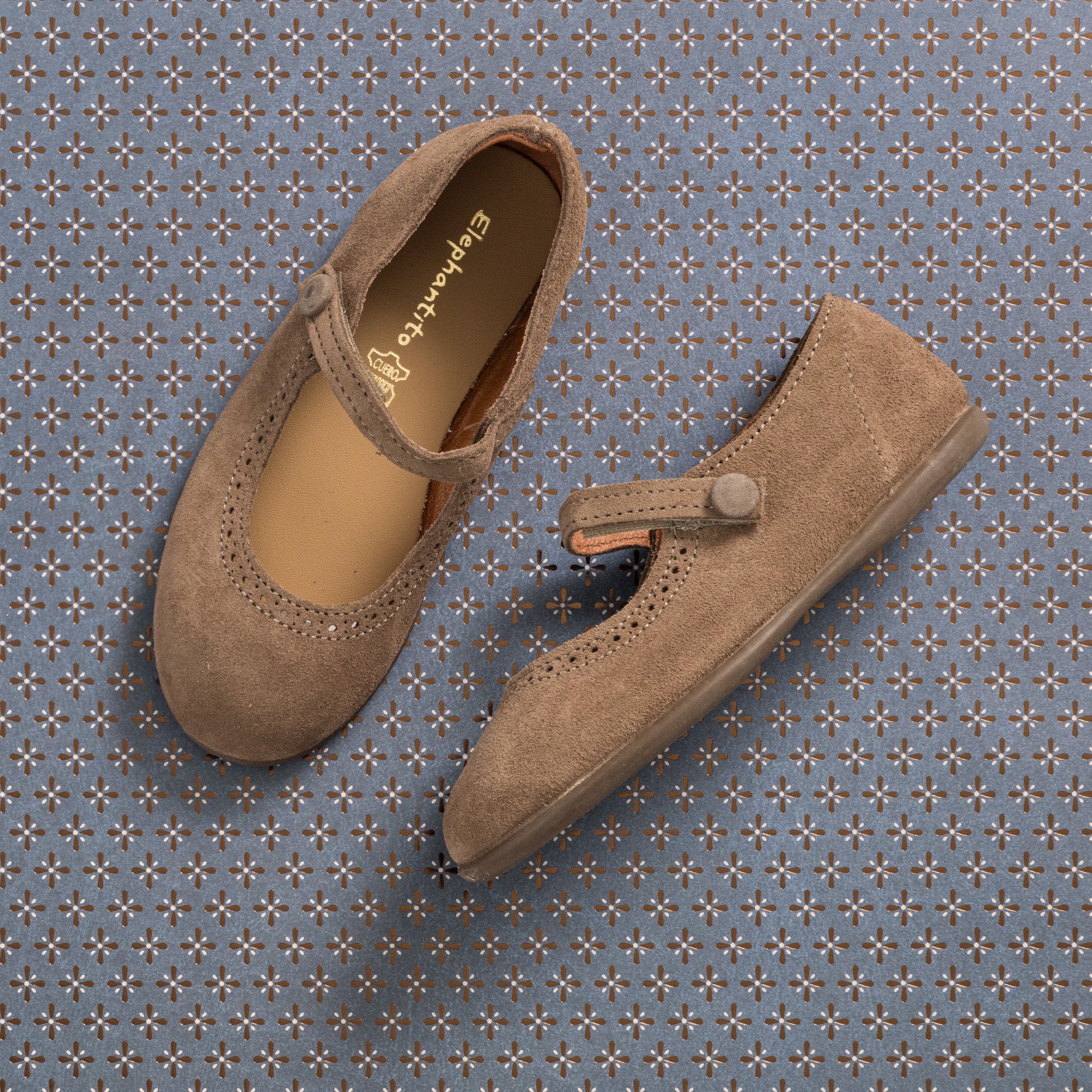 Beige suede toddler Mary Jane shoes with rounded toe and buttoned strap on patterned background.