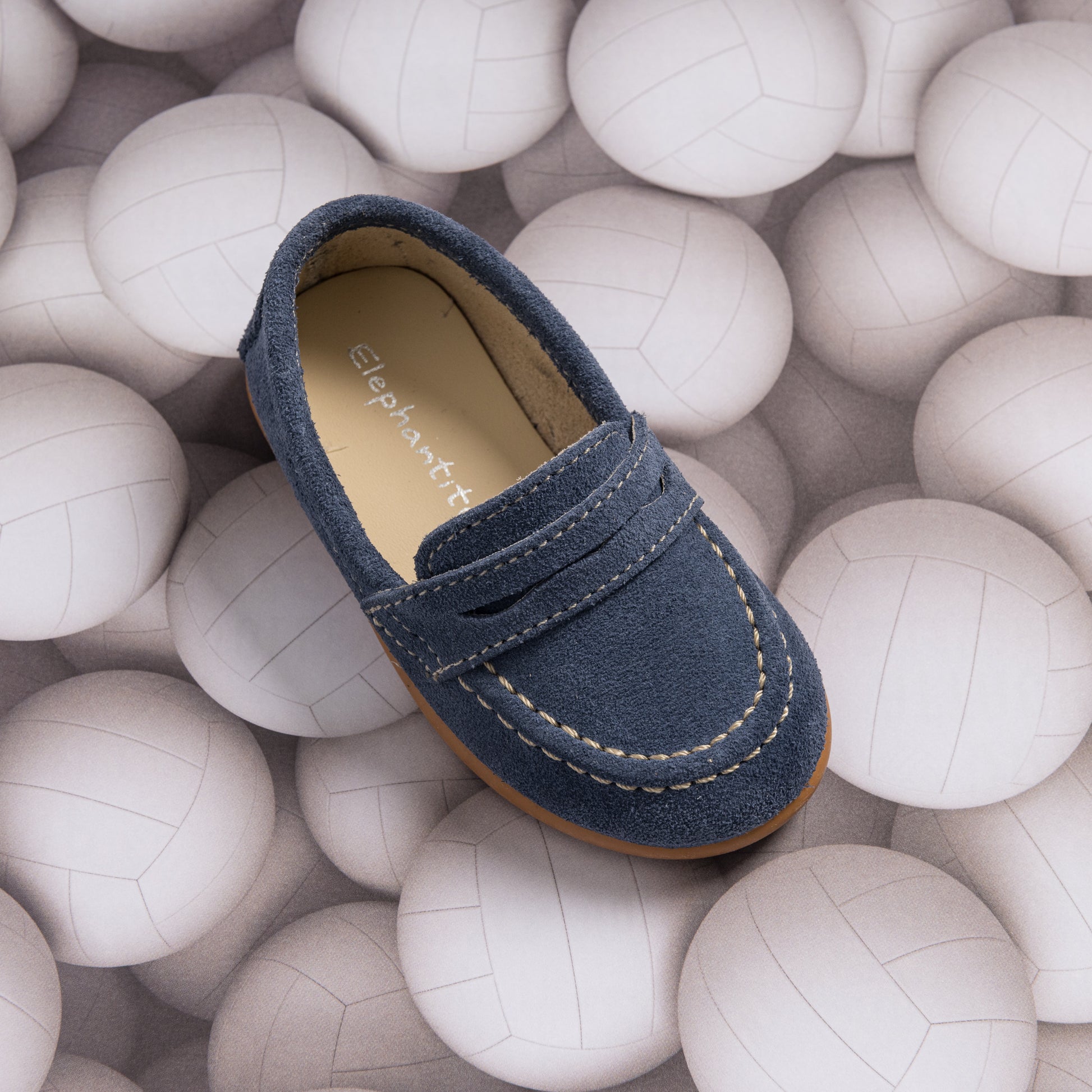 Navy blue suede penny loafer for toddlers with white stitching and brown sole on volleyball background.