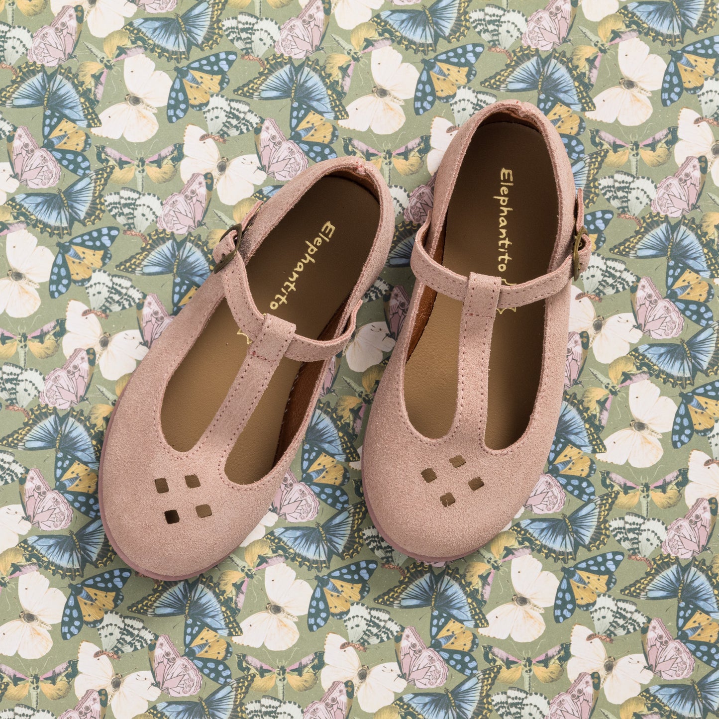 Pink suede T-bar toddler shoes with cutout details and buckle straps on floral background.