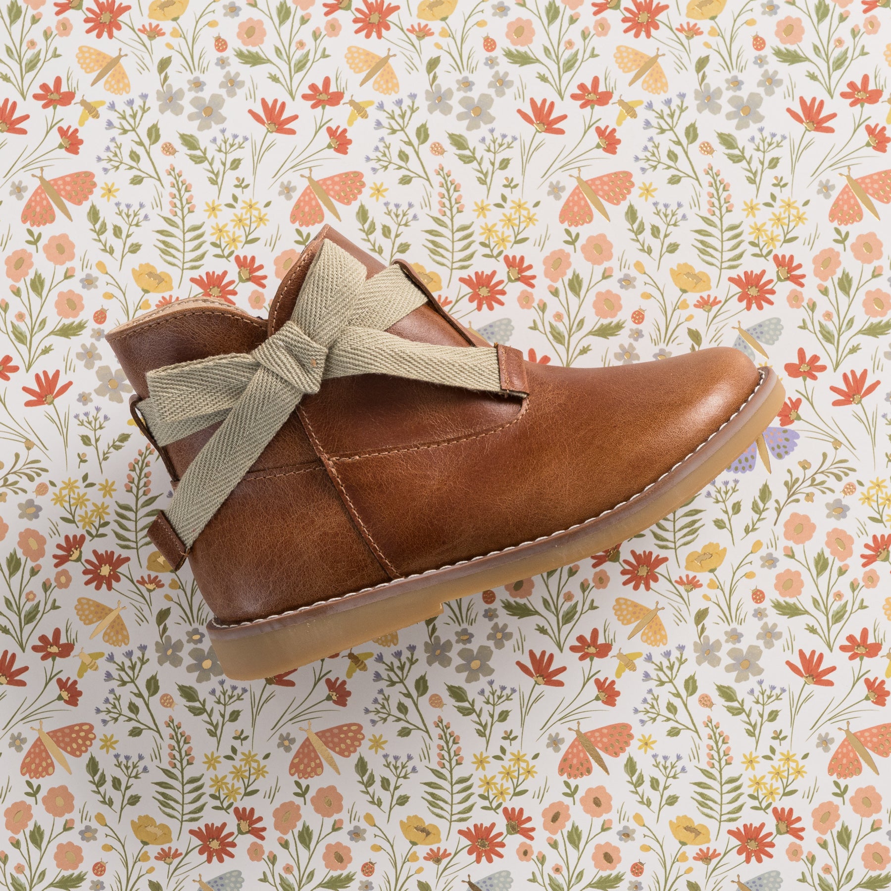 Brown leather toddler bootie with beige fabric bow and rubber sole on floral background.