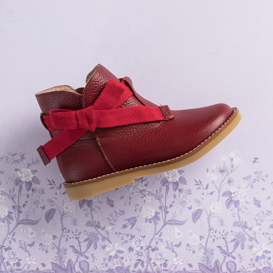Red leather toddler bootie with side bow and tan rubber sole on floral background.