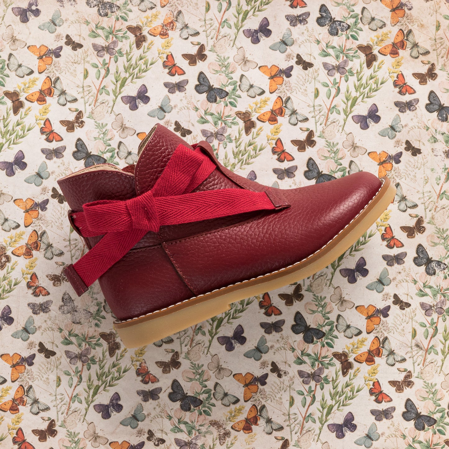 Red toddler bootie with large fabric bow and tan rubber sole on butterfly print background.