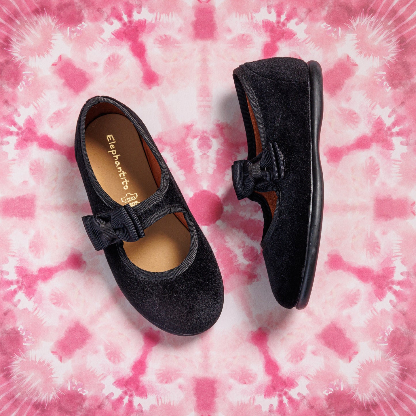 Black velvet girls Mary Jane shoes with a bow detail and rounded toe on tie-dye pink background.