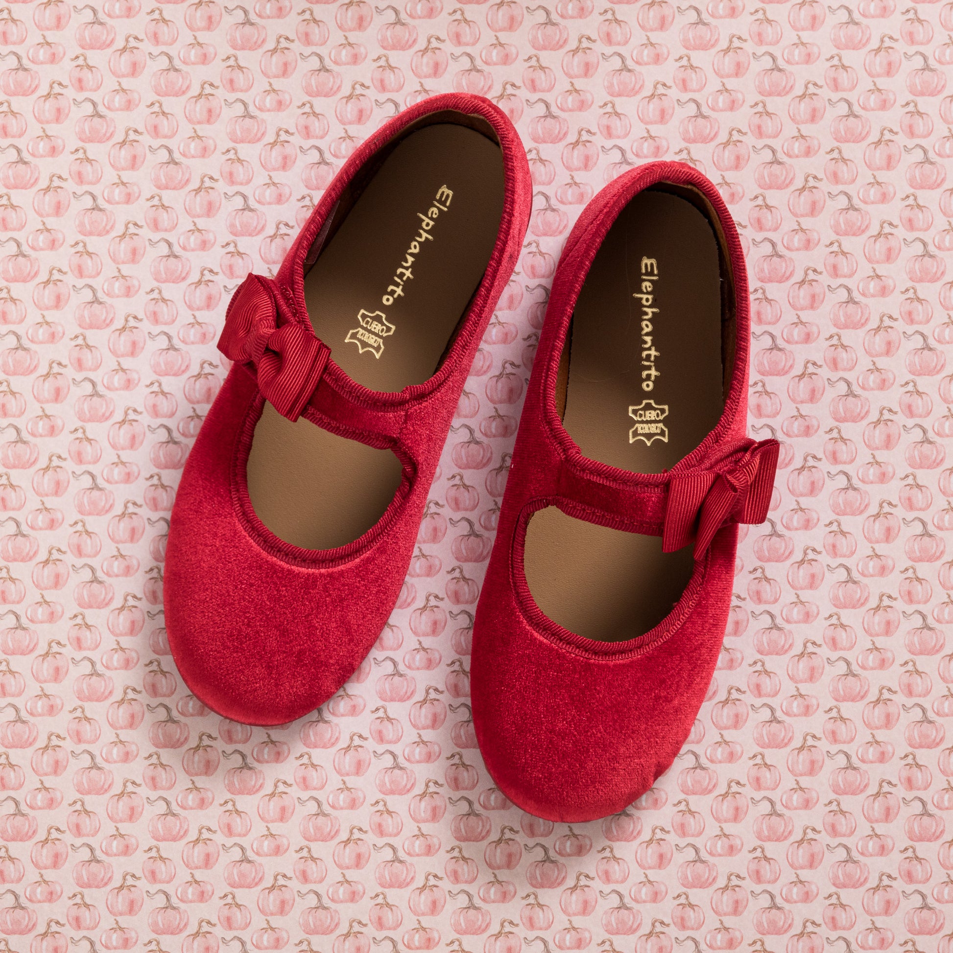 Red velvet Mary Jane shoes with side bows and rounded toes on pink patterned background.