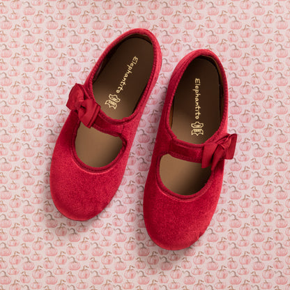 Red velvet Mary Jane shoes with side bows and rounded toes on pink patterned background.