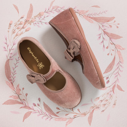 Pink velvet Mary Jane shoes with bow detail and rubber sole, displayed on floral background.