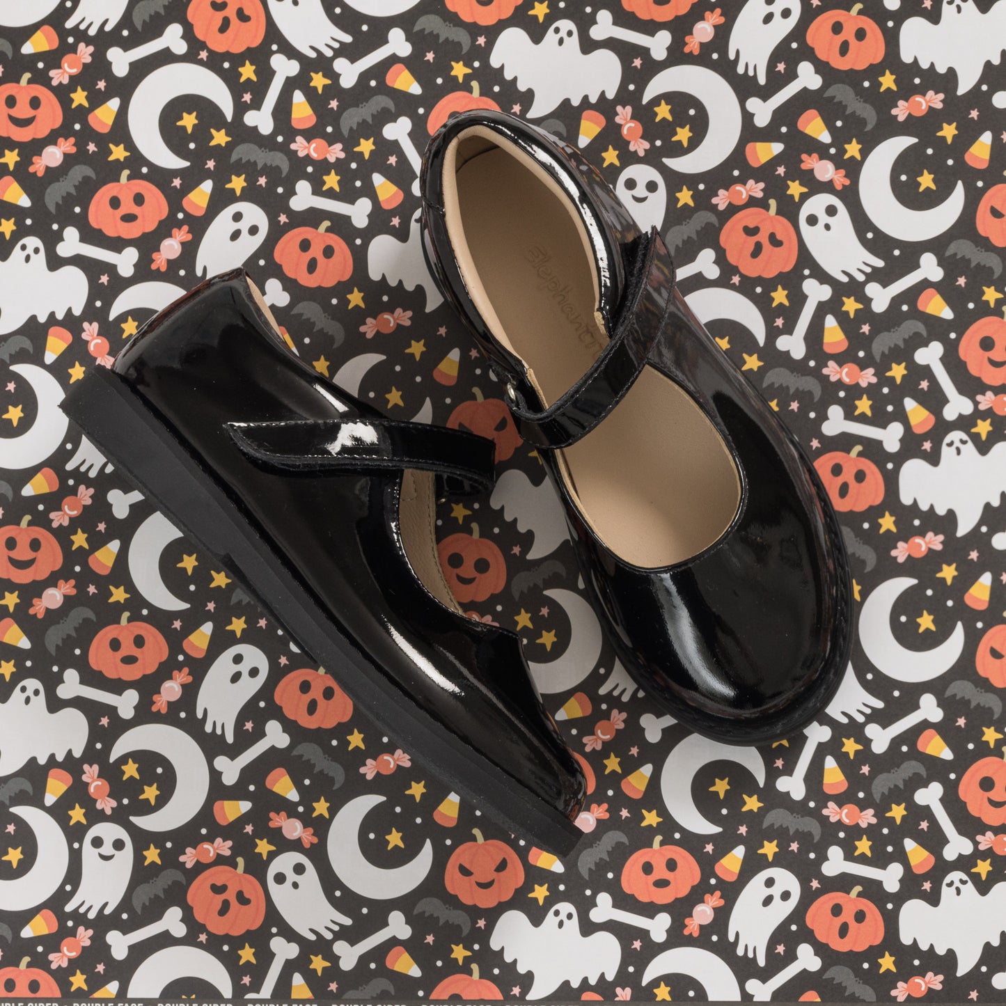 Black patent leather girls Mary Jane shoes with a strap and rounded toe on Halloween-themed background.