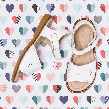 White children's sandal with scalloped edges and cutout detailing, tan sole on heart pattern background.