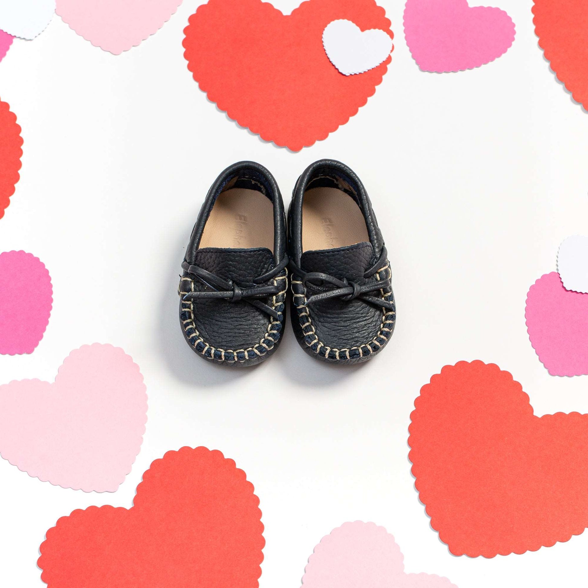 Navy blue baby driver loafers with bow detail and contrast stitching on white background with heart shapes.