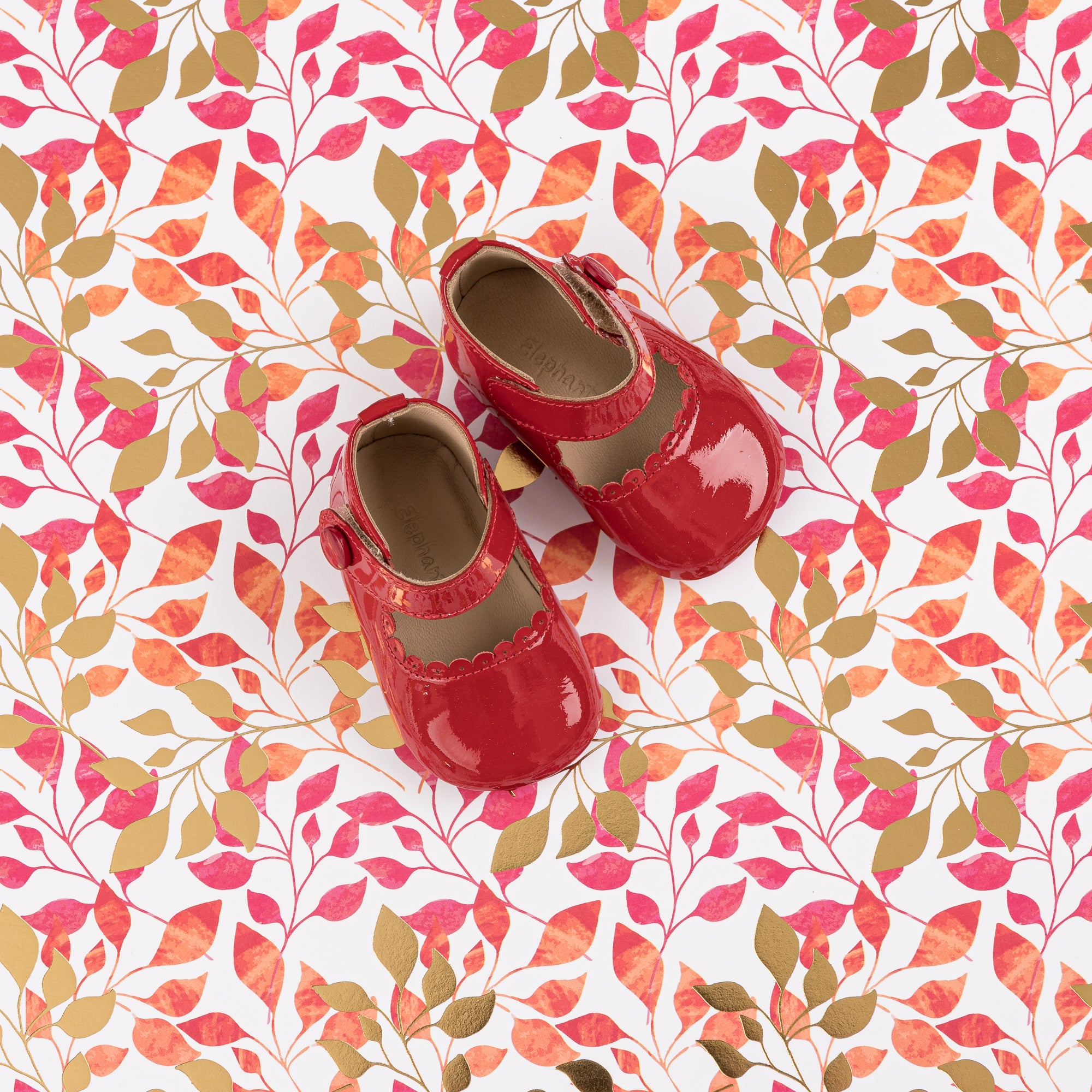 Glossy red patent leather Mary Jane baby shoes with velcro strap on colorful leaf-patterned fabric background.