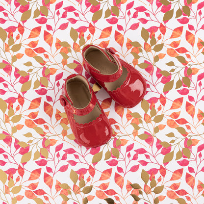 Glossy red patent leather Mary Jane baby shoes with velcro strap on colorful leaf-patterned fabric background.
