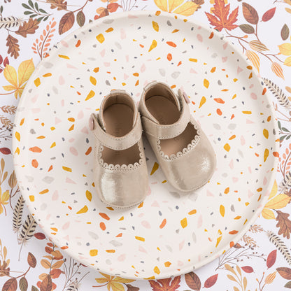 Beige toddler Mary Jane shoes with scalloped trim and Velcro strap on a white plate with autumn leaf print.