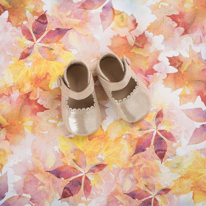 Beige baby Mary Jane shoes with scalloped edges and velcro straps on a colorful autumn leaf background.