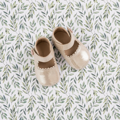 Cream-colored baby Mary Jane shoes with scalloped edges and soft leather material on a leafy patterned background.