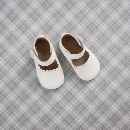 White baby Mary Jane shoes with scalloped edge and Velcro strap on gray plaid background.