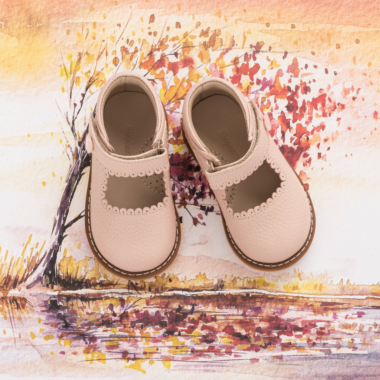 Pink leather Mary Jane toddler shoes with scalloped edges and velcro strap over printed autumn tree background.