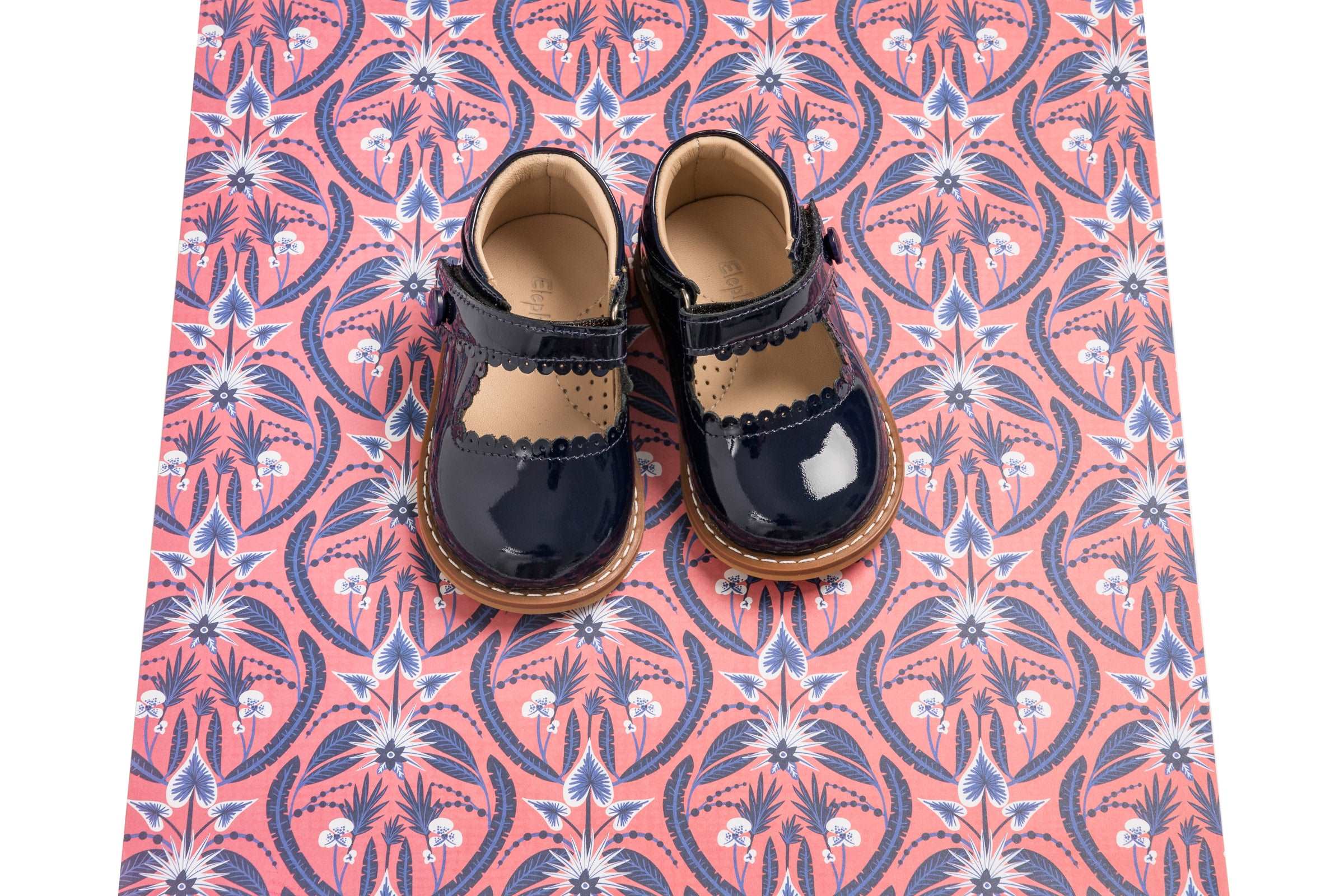 Toddler navy blue patent leather Mary Jane shoes with scalloped edge and tan sole on pink patterned fabric.
