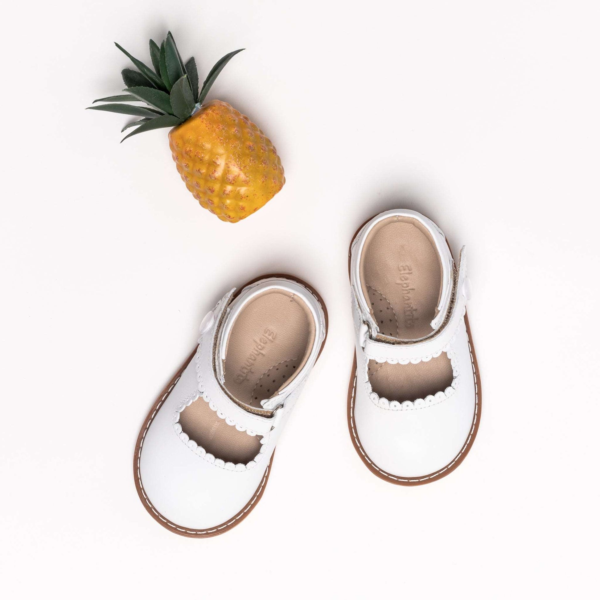 White toddler Mary Jane shoes with scalloped edges and tan soles, paired with a small pineapple accessory.