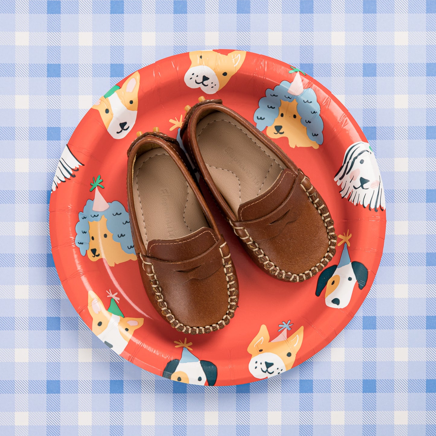 Natural brown baby moccasins with stitched detailing on a red plate featuring cartoon dogs.