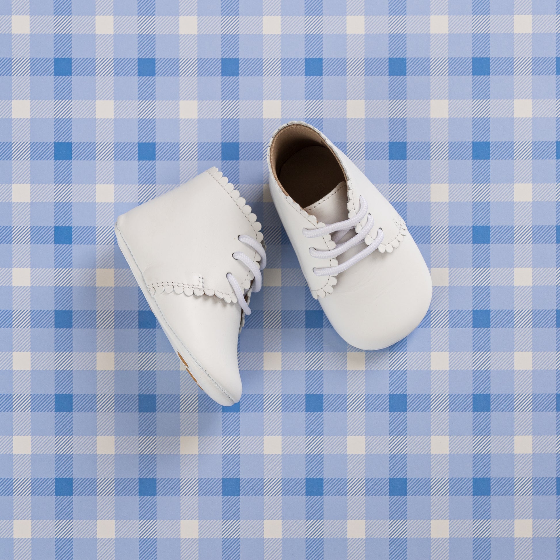 White baby booties with scalloped edges and lace-up fronts on a blue plaid background.