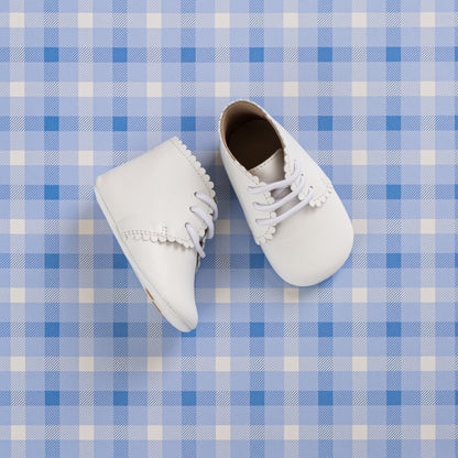 White baby booties with scalloped edges and lace-up fronts on a blue plaid background.