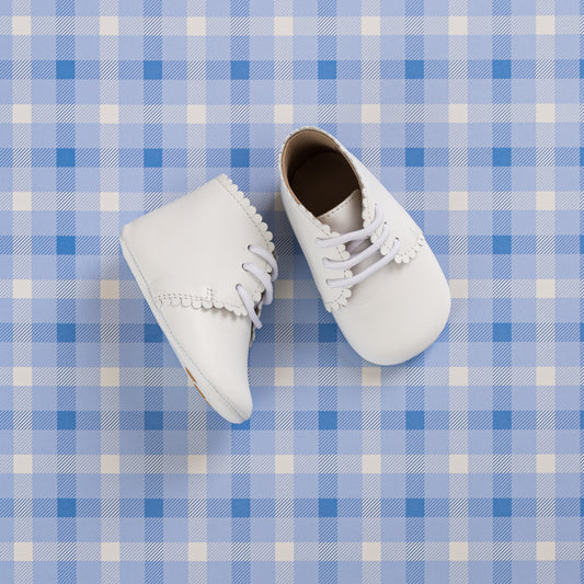 White baby booties with scalloped edges and lace-up fronts on a blue plaid background.