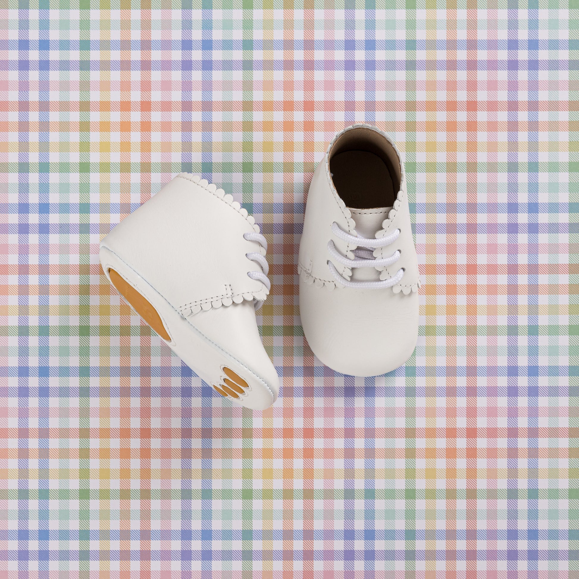 White infant booties with scalloped edges and lace-up design on pastel plaid background.