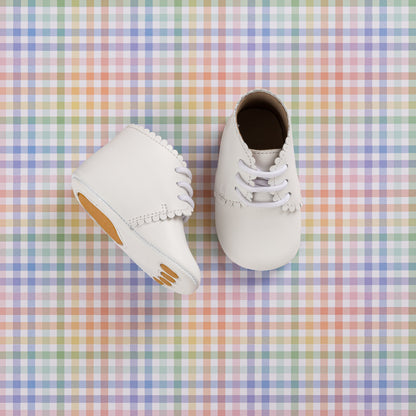 White infant booties with scalloped edges and lace-up design on pastel plaid background.